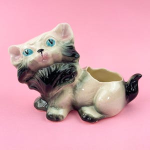 May include: A ceramic cat planter in a seated position, with a cream, gray, and black color scheme. The cat has blue eyes and a small opening on its back for planting. The planter is on a pink background.