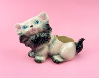 May include: A ceramic cat planter in a seated position, with a cream, grey, and black colour scheme. The cat has blue eyes and a small opening on its back for planting. The planter is on a pink background.