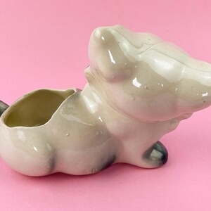 May include: A vintage ceramic planter in the shape of a cat, with a cream-colored body and black accents on the tail and paws. The planter has an open bowl for holding plants and sits on a pink background.
