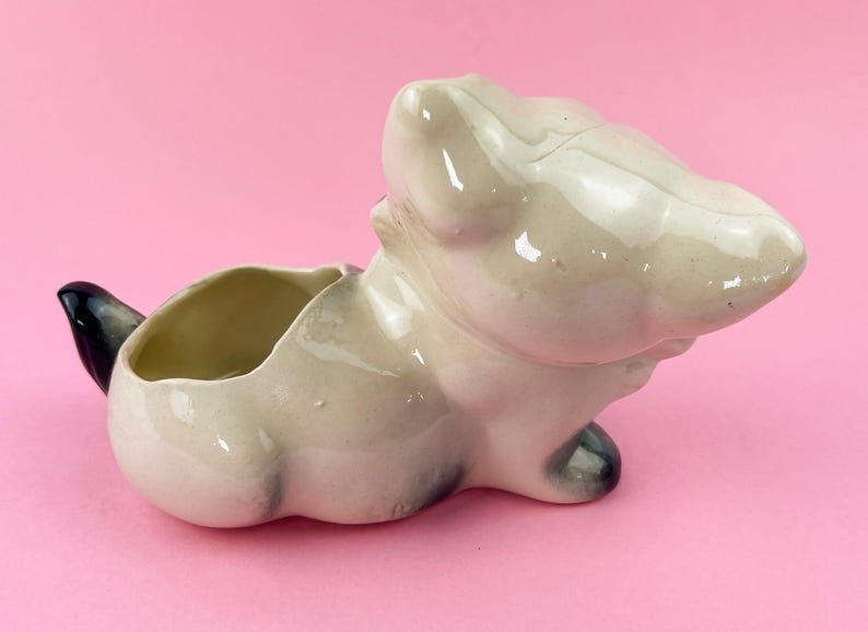 May include: A vintage ceramic planter in the shape of a cat, with a cream-colored body and black accents on the tail and paws. The planter has an open bowl for holding plants and sits on a pink background.
