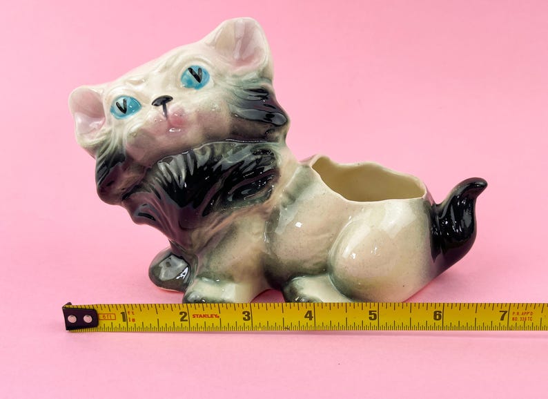 May include: A vintage ceramic cat planter with blue eyes, black and gray fur, and a pink nose. The planter is approximately 7 inches long. The cat is in a seated position with a hollowed-out back for plants.