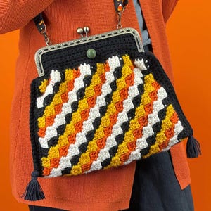 May include: A black crochet purse with a metal clasp and a candy corn pattern in white, orange, and yellow. The purse has a black frame, a green stone clasp, and a black tassel on each side. The strap has a candy corn design.