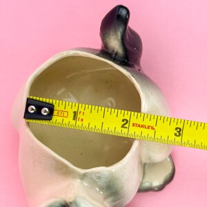 May include: A vintage ceramic planter in the shape of a dog, with a gray and white color scheme. The planter is approximately 4 inches long. The interior is beige, and the background is pink.