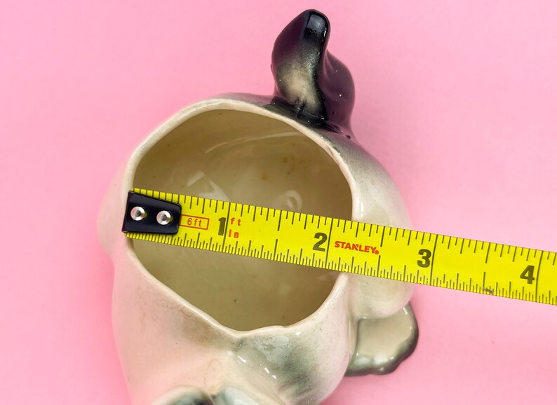 May include: A vintage ceramic planter in the shape of a dog, with a gray and white color scheme. The planter is approximately 4 inches long. The interior is beige, and the background is pink.