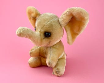 May include: A small, plush, tan elephant toy with large ears and black eyes. The elephant is sitting on a pink background.