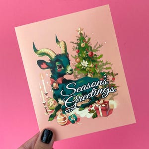May include: A Christmas card featuring a green and black goat with a red bow sitting under a Christmas tree with the text "Season's Greetings".