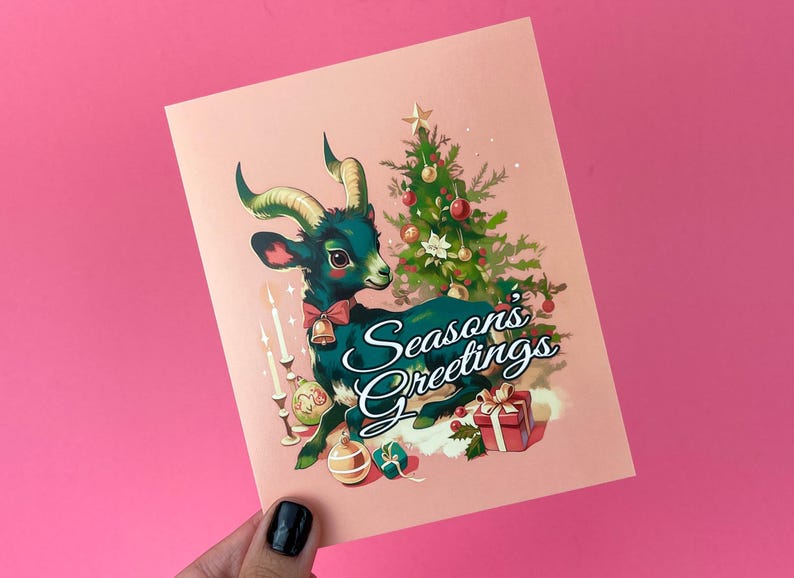 May include: A Christmas card featuring a green and black goat with a red bow sitting under a Christmas tree with the text "Season's Greetings".