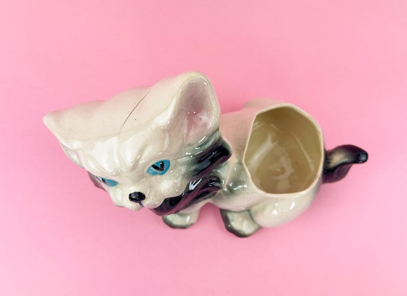 May include: A vintage ceramic cat planter in cream, gray, and black, with blue eyes. The cat-shaped planter has an open compartment on its back for holding small plants or decorative items. The planter is on a pink background.