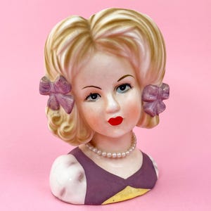 May include: A vintage ceramic bust of a woman with blonde hair styled in curls and adorned with purple bows. She has blue eyes, red lips, and a pearl necklace. The bust is wearing a purple and yellow dress.