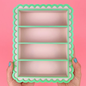 May include: A rectangular, four-shelf display shelf with a scalloped edge. The shelf is mint green with a light pink interior. The shelf is held by hands against a pink background.