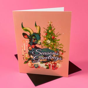 May include: A Christmas card featuring a blue and green goat with a red bow, sitting in front of a decorated Christmas tree. The card has a pink background and the text "Season's Greetings".