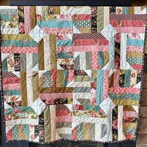 May include: A patchwork quilt with a variety of floral and striped patterns in shades of pink, teal, cream, and brown. The quilt is made of rectangular and triangular fabric pieces, creating a geometric design. The quilt is hanging on a wall.