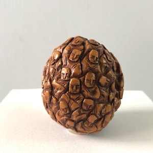 Może przedstawiać: Szczegółowa, brązowa rzeźba z drewna w kształcie kuli pokrytej wyrzeźbionymi twarzami. Twarze są stylizowane, z prostymi cechami i są ściśle upakowane, tworząc teksturowaną powierzchnię. Rzeźba jest ustawiona na białym tle.