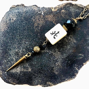 Op de afbeelding: Een ketting met een hanger in de vorm van een zwarte en witte mahjong-tegel, een zwarte kristallen kraal en een gouden spijkerbedel. De ketting ligt op een donkergrijze achtergrond.