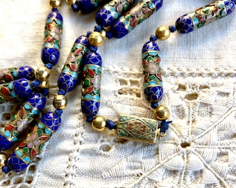 Puede incluir: Un collar de múltiples hilos con cuentas de esmalte azul, verde y rosa. Las cuentas están decoradas con motivos florales y están separadas por separadores dorados. El collar tiene un cierre dorado.