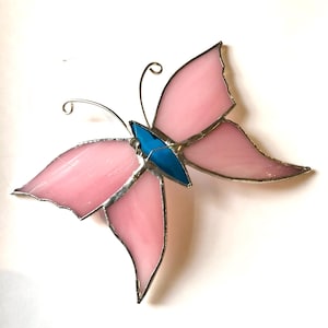 Puede incluir: Una mariposa de vidriera con alas rosas y cuerpo azul. Las alas están delineadas en plata y tienen una apariencia translúcida. La mariposa tiene delicadas antenas de alambre y está sobre un fondo blanco.