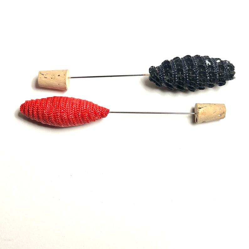 Puede incluir: Dos alfileres decorativos con tapas de corcho y cabezas de tela de diferentes colores. Un alfiler tiene una cabeza de tela roja y el otro tiene una cabeza de tela negra.