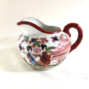 Puede incluir: Una jarra de crema de porcelana blanca con un asa roja. La jarra está decorada con un diseño de inspiración japonesa que presenta mujeres con ropa tradicional, flores y una pagoda.
