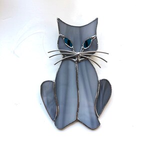 May include: A gray stained glass cat with blue eyes. The cat is sitting with its legs tucked under its body.