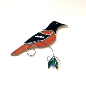 Puede incluir: Un pájaro de vidrio tintado con alas negras y naranjas, posado en un tallo verde con una cuenta azul.