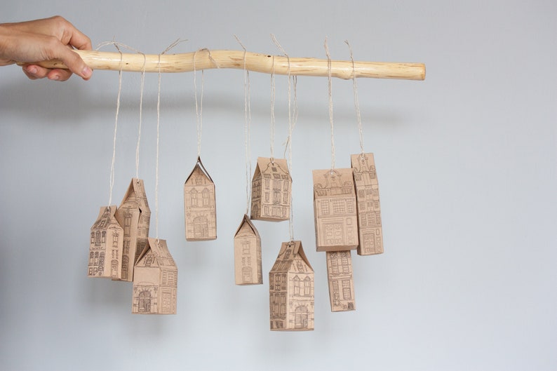 May include: A wooden branch with brown paper houses hanging from it with twine. The houses are decorated with a black and white print of a building.