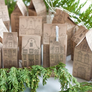 May include: A set of small, brown paper boxes shaped like houses with black and white illustrations. The boxes are arranged in a row on a white surface with green foliage in the foreground.
