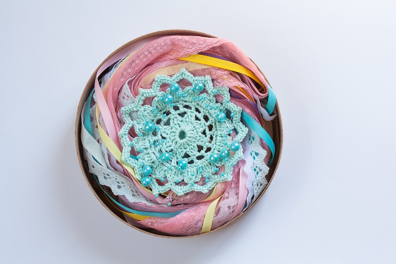 May include: A round container filled with a variety of pastel colored ribbons, lace, and a crocheted doily with turquoise beads.
