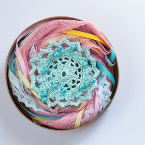 May include: A round container filled with a variety of pastel colored ribbons, lace, and a crocheted doily with turquoise beads.