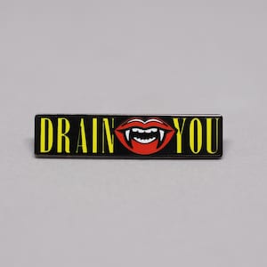 May include: A rectangular black enamel pin with the words "DRAIN YOU" in yellow, and a red vampire mouth graphic with white fangs. The pin is set against a gray background.