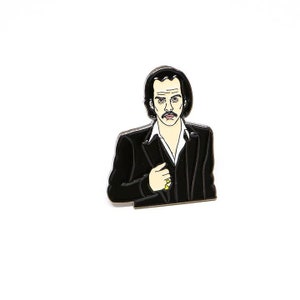 Puede incluir: Pin de esmalte negro y blanco de un hombre con cabello oscuro y bigote que lleva una chaqueta de traje negra. Está mirando hacia un lado y tiene una expresión seria.