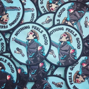 May include: A collection of round, blue embroidered patches with the text "Nothing Feels Good" in white. The patches feature a stylized image of a person with a distressed look on their face.