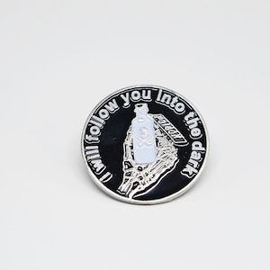 May include: A black and silver enamel pin featuring a skeleton hand holding a bottle labelled "Poison". The text surrounding the image reads "I will follow you into the dark."