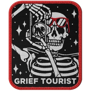 Puede incluir: Un parche bordado en blanco y negro con un esqueleto que lleva gafas de sol rojas y sostiene una cámara. El texto "GRIEF TOURIST" está impreso debajo del esqueleto.