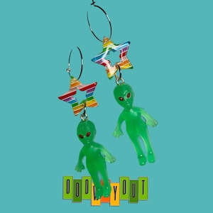 Op de afbeelding: Een paar zilveren creolen met regenboogsterrencharms en groene alienfiguren die eraan hangen. De oorbellen zijn tegen een turquoise achtergrond met de tekst "ODDLY OUT" in gele en groene letters.