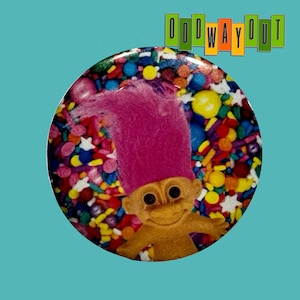 May include: A round button featuring a Troll doll with a pink, upright hair. The background is a colorful mix of sprinkles and candies. The text "ODD WAY OUT" is at the top in a rectangular font.