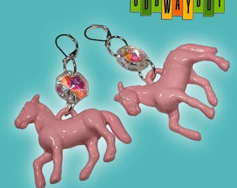 May include: A pair of pink horse earrings with silver-tone hooks and iridescent crystal accents. The earrings feature a small, pink horse charm and are set against a teal background. The text "ODD WAY OUT" is in the upper right corner.