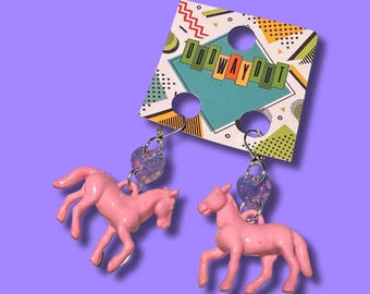 May include: Pink plastic horse earrings with a purple glitter heart charm. The earrings are on a white card with the text "Odd Way Out" in rainbow colors.