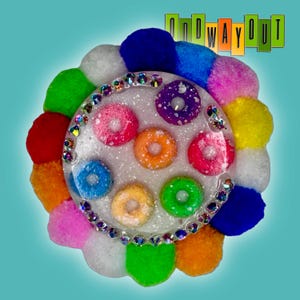 Puede incluir: Una pieza circular de resina con una base blanca brillante incrustada con seis cereales coloridos en forma de anillo. El perímetro está bordeado por pequeños diamantes de imitación transparentes y rodeado por pompones difusos multicolores. El texto dice ONDWAYOUT.