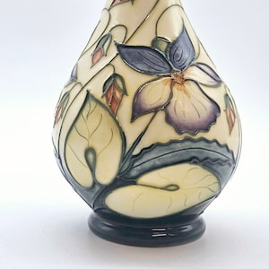 May include: A cream-colored ceramic vase with a dark blue rim and base. The vase is decorated with a floral design featuring purple flowers, green leaves, and orange buds.