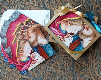May include: A set of blank greeting cards with a watercolor painting of a holy family. The painting depicts a mother, father, and baby in a loving embrace. The cards are packaged in a brown box with a gold ribbon.