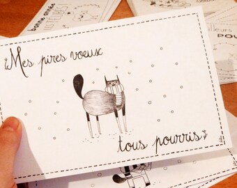 May include: A white greeting card with a black cat illustration wearing a scarf, surrounded by falling snow. The card has the handwritten text "Mes pires voeux tous pourris" in black ink, with a dashed border.