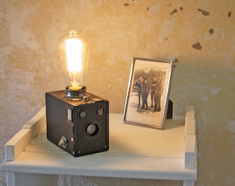 Könnte beinhalten: Eine schwarze Vintage-Kamera, die zu einer Lampe umfunktioniert wurde, mit einer großen Edison-Glühbirne. Die Lampe steht auf einem weißen Regal neben einem gerahmten Schwarz-Weiß-Foto. Der Hintergrund ist eine strukturierte, hellfarbige Wand.