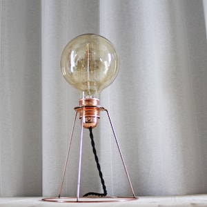 May include: A copper-colored desk lamp with a large, round, amber-toned light bulb. The lamp has a minimalist design with a circular base and three supporting legs. A black, braided cord extends from the bulb. The lamp is approximately 12 inches tall.