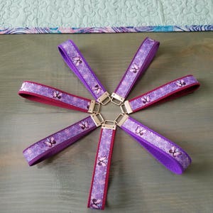 Op de afbeelding: Zes paarse en bordeauxrode sleutelhangers met een vlinderpatroon. De sleutelhangers zijn in de vorm van een ster op een houten oppervlak gerangschikt.