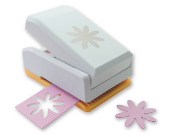 May include: A white paper punch with a silver daisy design on the top. The punch is open and a pink paper with a daisy cut out is visible. A pink daisy cut out is also visible on the table.