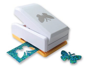 May include: A white butterfly punch with a yellow base. The punch is being used to cut out a butterfly shape from a piece of blue and green glitter paper. A cut-out butterfly is shown to the right of the punch.