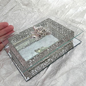May include: A rectangular glass jewellery box featuring a mirrored base and a silver-toned metal filigree frame. The lid is topped with a decorative silver-toned metal rose ornament. The box has a reflective surface and intricate cut-out metal detailing.