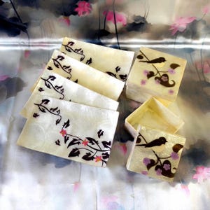 May include: A set of rectangular mother-of-pearl trays and a square lidded box. The items feature a cream-coloured iridescent surface decorated with black vine silhouettes, small pink flowers, and a black bird perched on a flowering branch.