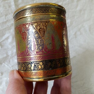 May include: A small, round, brass container with intricate floral designs in red, green, and gold. The container has a lid and is decorated with a black band.