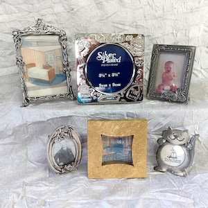 May include: A collection of five photo frames including a tall ornate silver frame, a square silver-plated frame with baby motifs, a rectangular silver frame, an oval silver frame, a square gold-textured frame, and a silver cat-shaped frame.